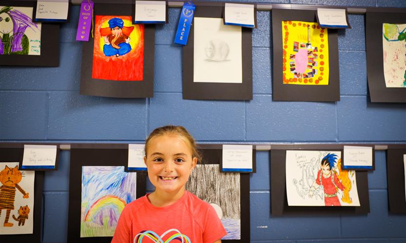 A girl wearing a coral shirt smiles while standing beneath student artwork